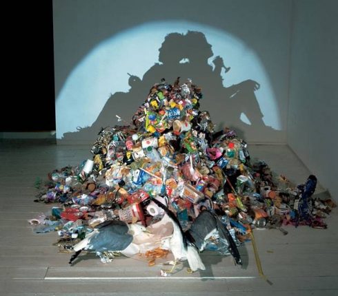 A large pile of trash lends to a shadow of two people sitting back to back leaning on each other