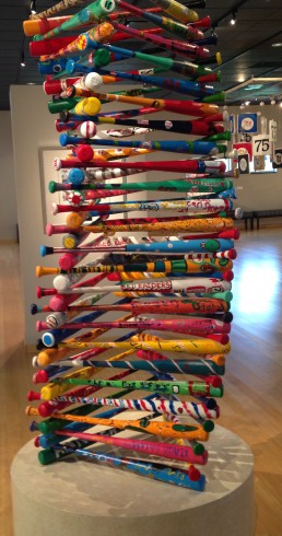 Image of a large sculpture constructed of colorfully painted baseball bats stacked up into a large 3D triangular shape.