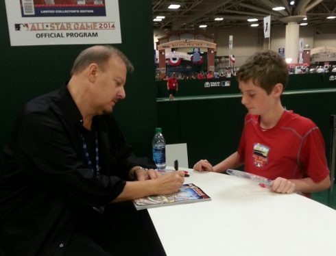 Charles Fazzino sitting at a table at the MLB All Star Fan Fest signing a program for a young boy.