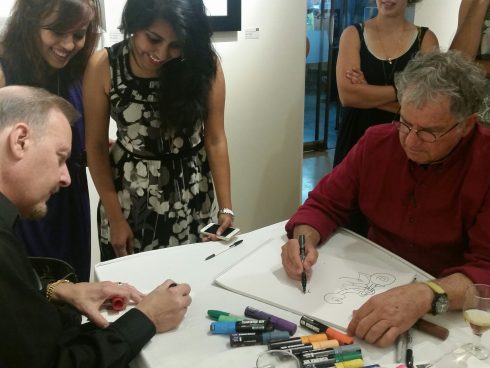 Charles Fazzino and David Gerstein working on drawings while onlookers smile with intrigue