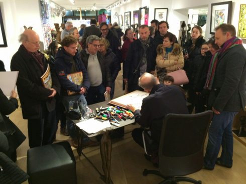 A group of individuals surrounding Charles Fazzino who is sitting down drawing at an Art Gallery