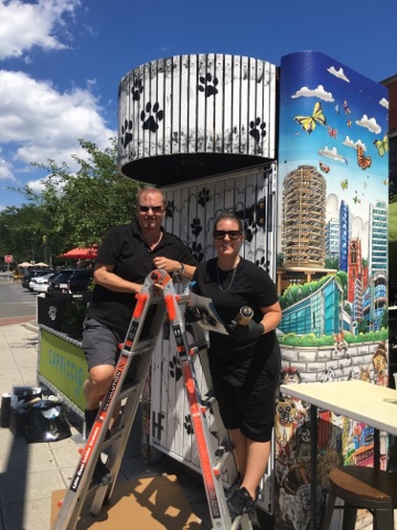 Charles and Heather Fazzino standing on an orange ladder in front of their latest pop-art piece, Downtown Dogs in Stamford