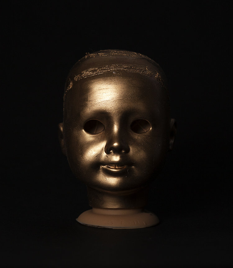 Gold painted doll head on a black backdrop shot by photographer Nicholas Rouke
