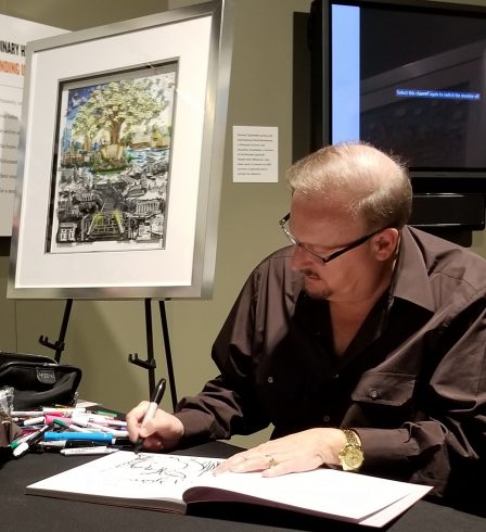 Charles Fazzino sitting at a black table, signing books in front of his 3D Pop art piece "After the Darkness" at the The Holocaust Memorial and Tolerance Center of Nassau County