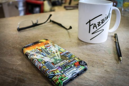 Fazzino pop art iPhone case next to a Fazzino logo mug and a pair of glasses on a wood table. 