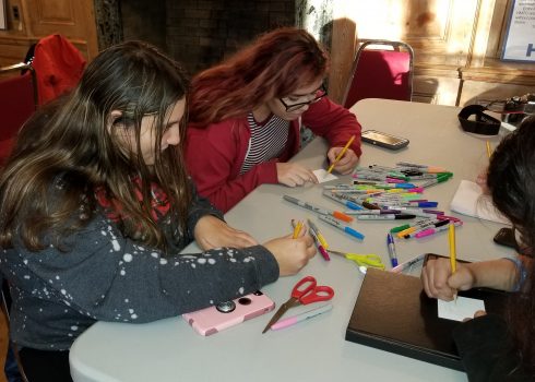 Two young women sitting at a white fold up table, surrounded by multi-colored sharpies, using pencils to sketch out 3d pieces for Charles Fazzino's recreation of "After the Darkness" pop art piece