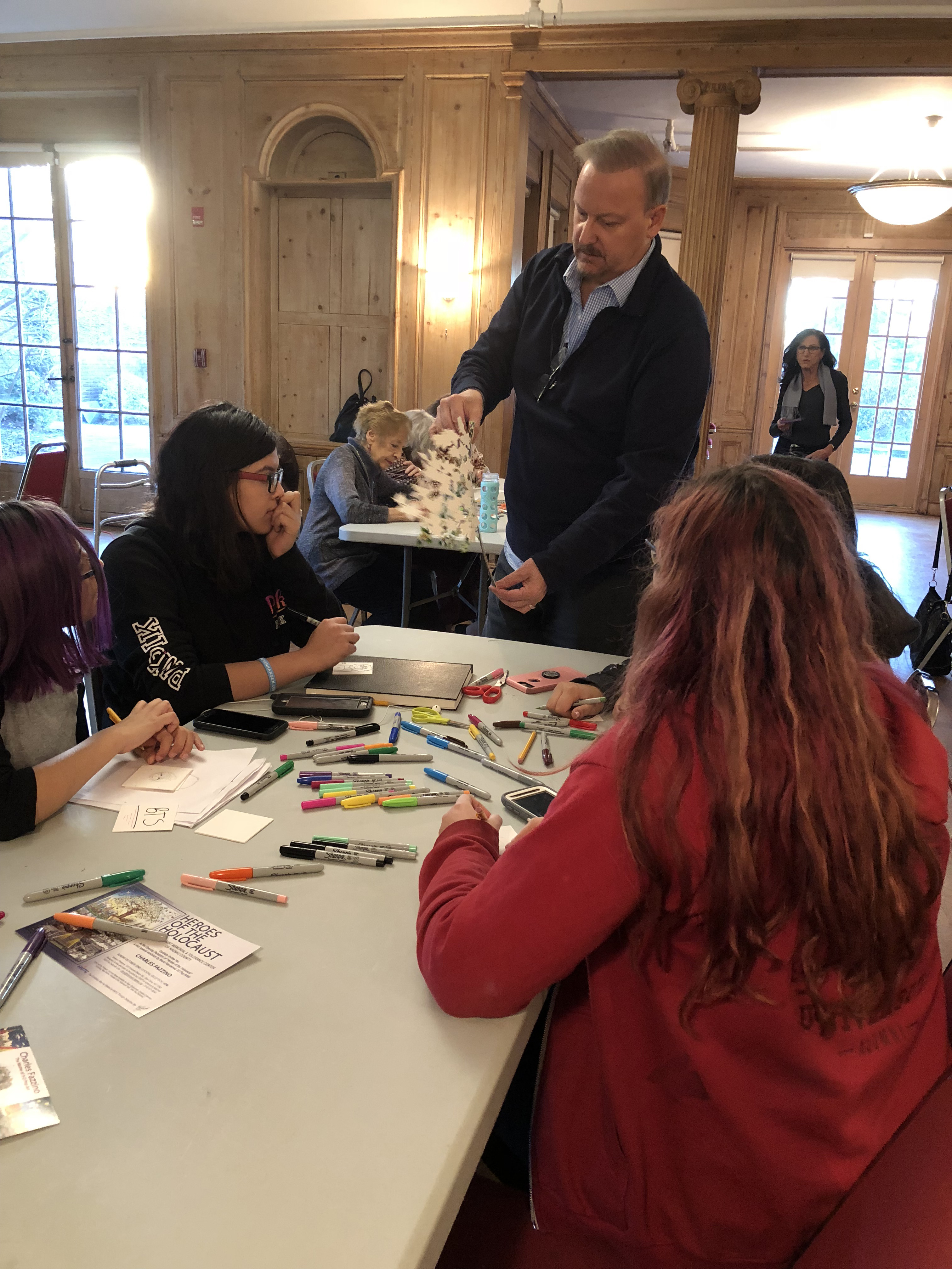 Charles Fazzino holding up the tree as young adults sit at a table and watch how he creates his 3D pop art work. 