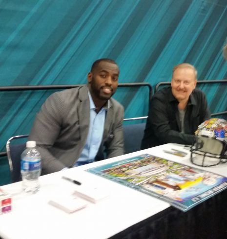 Fazzino and NFL players sit at a table signing posters during the Super Bowl