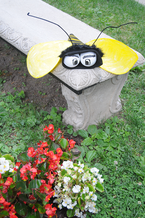 A felt bee with baseballs for eyes perched on top of a stone bench in front of flowers