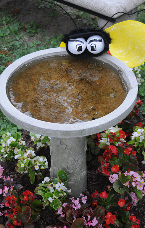 A bee made out of felt with baseballs for eyes perched on top of a bird bath with flowers in front