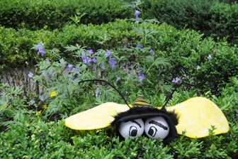 A felt bee with baseballs for eye perched on top of a green hedge with purple flowers in the background