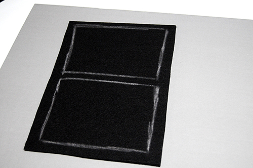 A piece of black felt on a grey paper board with chalk drawings to mark out a window