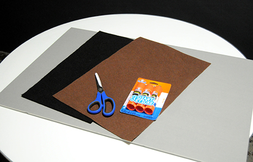 Grey paper board, brown and black felt, scissors and glue stick supplies on a white table