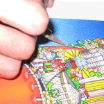 hand with exactoknife cutting a 3d pop art print - hand holding an pop art print of the new york city skyline in the shape of a red apple and a nyc subway train going through it like a worm, floating on an island next to the statue of liberty- 3D Pop Artist Charles Fazzino