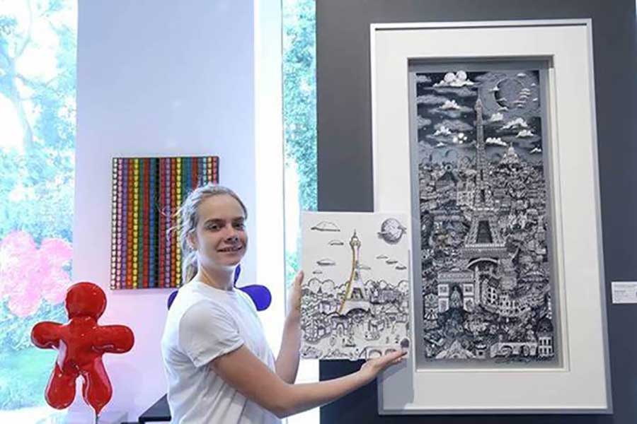 A student holds up artwork inspired by a Fazzino piece behind it from a virtual art workshop