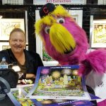 Charles Fazzino sits behind a table displaying art pieces, with a pink and yellow baseball mascot to his right