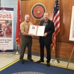 Charles Fazzino and Lt. Gen. Bellon stand in front of an American flag and US Marine Corps seal, holding a certificate