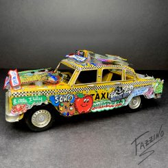 Side view of a yellow NYC taxi sculpture, featuring the words "Soho" and "Taxi" as well as a smiling red apple and other decorations on the hood and roof