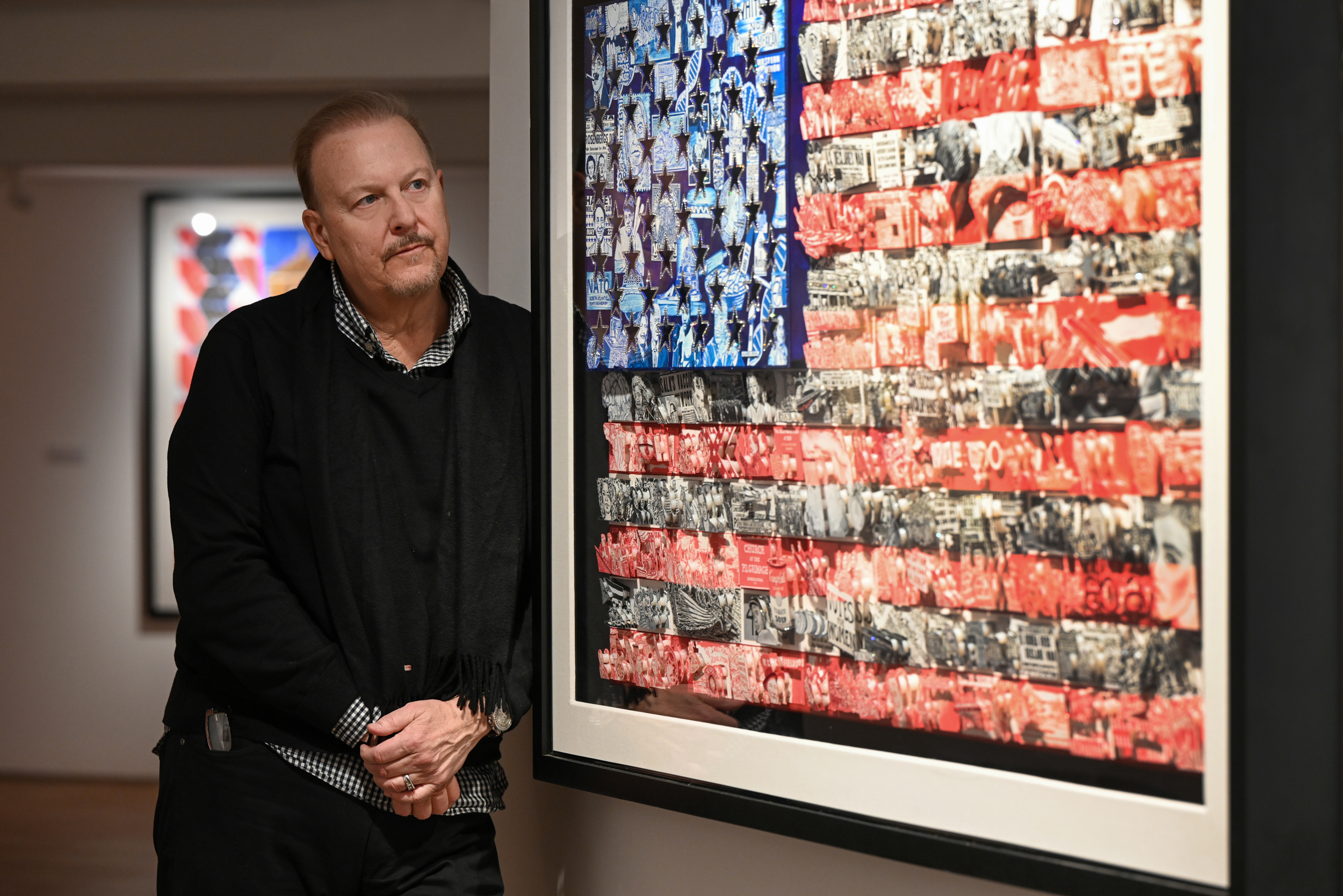 Fazzino se apoya en la pared de la galería junto a la bandera estadounidense de arte pop en 3D que presenta eventos históricos y figuras notables en azul, rojo y blanco.